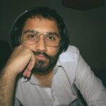 A thoughtful bearded man with glasses and headphones in a casual portrait setting.