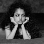 A thoughtful young woman with curly hair lies on a bed in a black and white portrait. Moody and introspective.