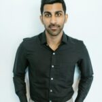 Portrait of a serious man in a black shirt with hands in pockets against a light background.